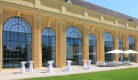 背景にアーチ型の窓のウィーン・オランジェリーがあり、庭園中庭にバーテーブルや休憩所が設置されています。シェーンブルン宮殿劇場管弦楽団のコンサートの前のアペリティフにぴった